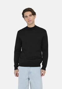 Maglione nero a maniche lunghe con un collo alto, polsini e fondo a coste, realizzato in un tessuto morbido ed elastico. Abbinato a jeans in denim azzurro chiaro.