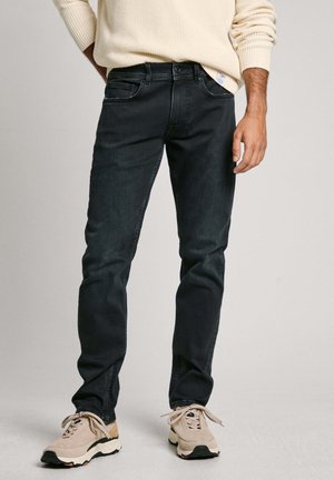 Jeans de mezclilla oscura con un corte ajustado, que cuentan con cinco bolsillos y una sutil textura desteñida. Combinados con zapatillas deportivas beige.