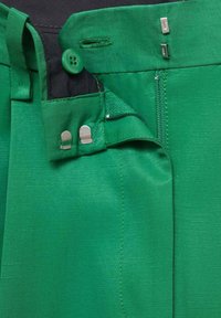 Pantalon en tissu vert avec bouton, fermeture éclair et agrafes métalliques, partiellement dézippé et déboutonné sur fond noir.