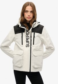 Weiße Jacke mit schwarzen Akzenten, mit Reißverschlusstaschen und einem auffälligen Logo auf der Vorderseite. Sie verfügt über eine Kapuze und geräumige Fronttaschen.