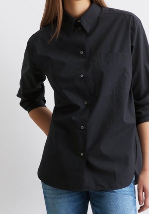 Kobieta w czarnej koszuli z długim rękawem zapinanej na guziki z kieszenią na piersi oraz w niebieskich dżinsach, na jasnym, jednolitym tle.