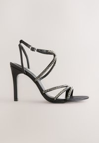 Črna sandaleta z visokim petom z več prečnimi trakovi, okrašenimi s prozornimi kristali. Gladek tekstura, odprti prst in nastavljiv trak okoli gležnja.