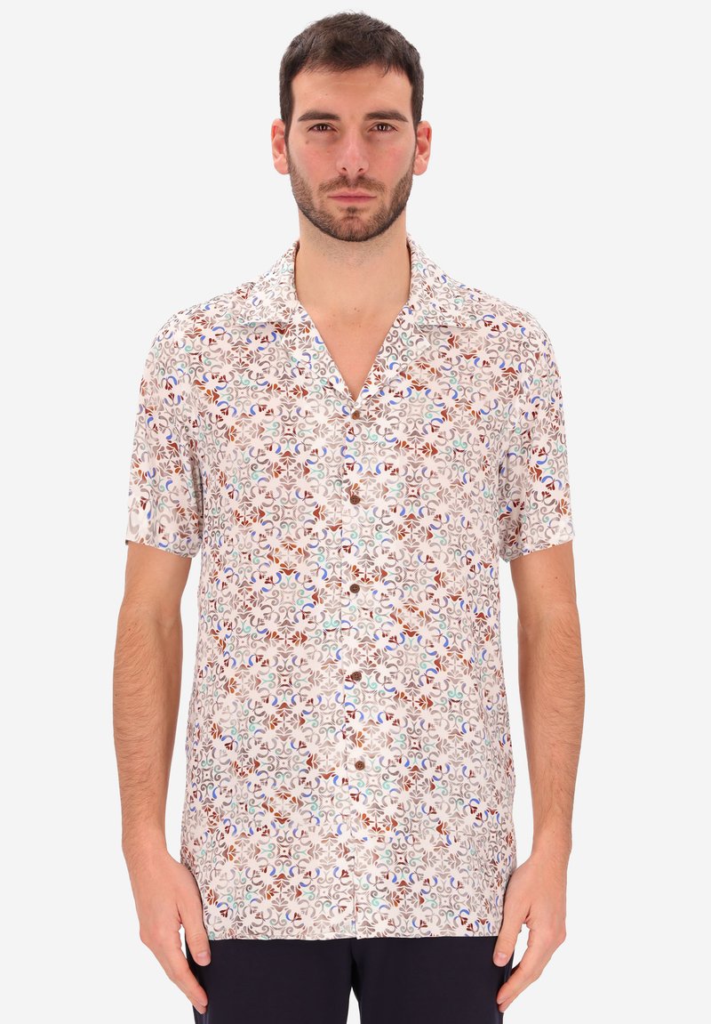 Uomo con camicia a maniche corte, botton-down, con un motivo astratto multicolore, in piedi contro uno sfondo bianco uniforme.