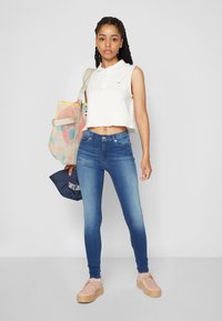 Bijela bezrukavna polo majica s skraćenim rubom, plave skinny traperice visokog struka i svijetlo ružičaste platforme. Model drži šarenu torbu.