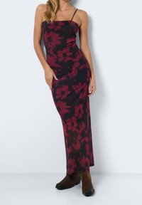 Frau in einem ärmellosen, schwarz-dunkelroten, floral gemusterten Maxikleid mit dünnen Trägern und braunen Stiefeletten, die vor einem schlichten Hintergrund steht.