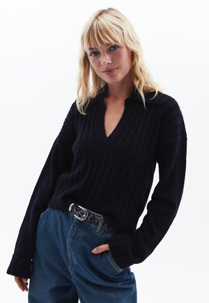 Navy gestreifter V-Ausschnitt-Pullover mit langen Ärmeln, der einen Kragen besitzt. Kombiniert mit hochtaillierten blauen Jeans und einem gemusterten Gürtel.