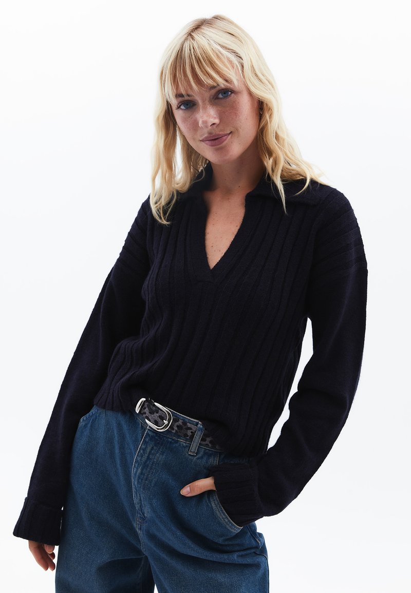 Maglione a coste blu navy con scollo a V e maniche lunghe, dotato di colletto. Abbinato a jeans in denim blu a vita alta e una cintura fantasia.