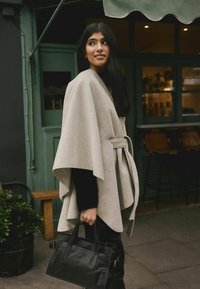 Heled hall villane kapuuts, millel on vöö, drapeeritud disain, kantud musta pluusi üle ja hoides struktureeritud musta nahk-käekotti.