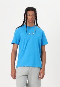 Modra bombažna majica s kratkimi rokavi in okroglim izrezom, na sprednji strani z majhnim srebrnim ovalnim logotipom. Nošena zivo sogrom z dolgimi hlačami.