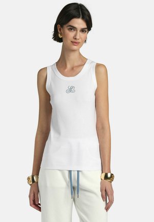 Žena nosi bijelu pamučnu majicu bez rukava s malim logom "LB" i svijetle hlače s plavim vezicama, ukrašena zlatnim narukvicama.