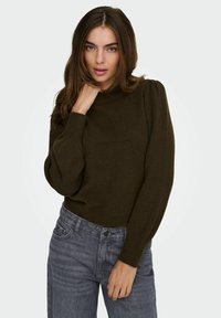 Jeune femme portant un pull à manches longues vert olive foncé avec des épaules bouffantes et un col montant, associé à un jean gris délavé, sur fond blanc.