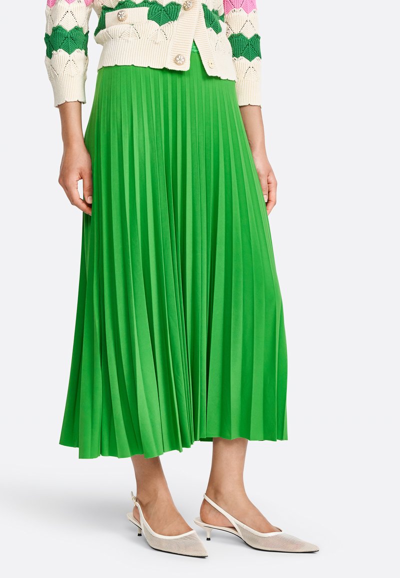 Grüne plissierte Midirock aus leichtem Stoff mit klaren Falten. Getragen mit weißen Slingback-Schuhen mit spitzem Absatz.
