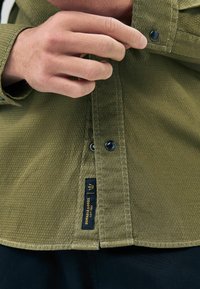 A hand buttons an olive green shirt with textured fabric, featuring a black and gold label reading "Durable Goods" near the hem.