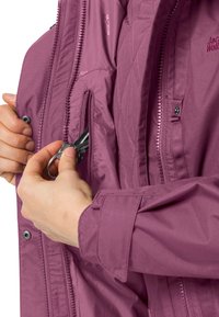 Des mains glissant un porte-clés avec des clés dans la poche intérieure zippée d'une veste violette Jack Wolfskin.