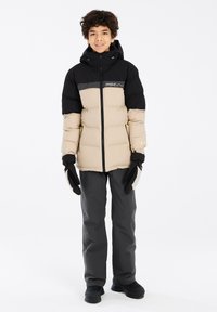 Giacca imbottita nera e beige con zip frontale, cappuccio e logo. Abbinata a pantaloni grigio scuro e guanti, tutti adatti per il freddo.