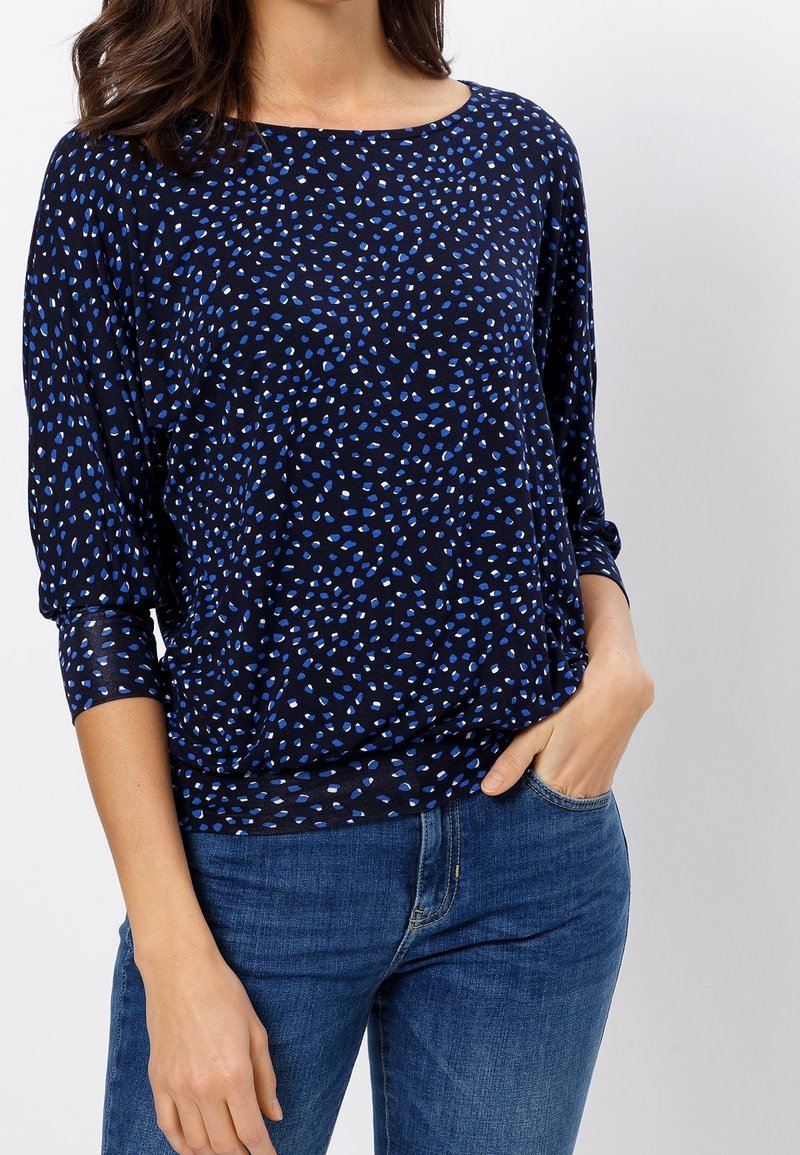 Femme portant un haut bleu marine avec un motif à pois bleu et blanc et un jean bleu, une main dans la poche, sur un fond uni.