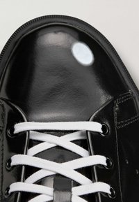 Crni tenisica od lakirane kože sa bijelim vezicama, okruglim vrhom i glatkom površinom. Ima udubice za vezice i zaobljenu petu.