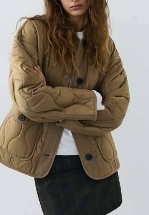 Femme portant une veste matelassée marron avec des boutons noirs et une jupe à carreaux foncée, bras croisés, debout devant un fond clair uni.