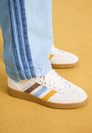 Białe sneakersy Adidas Spezial z niebieskimi, brązowymi i żółtymi paskami, noszone z jasnoniebieskimi dżinsami na żółtej podłodze.