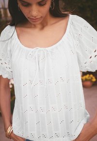 Blouse blanche à manches bouffantes, ornée de broderies ajourées et d'un décolleté à cordon. Texture douce et détails subtils en motif visibles.