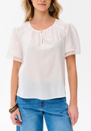 Kobieta w białej bluzce z krótkim rękawem, koronkowymi mankietami i dekoltem z kluczykiem, w połączeniu z niebieskimi dżinsami i czarnymi kolczykami z wzorem.