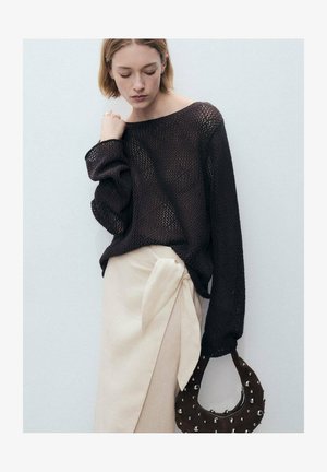 Pull en laine marron avec des manches surdimensionnées, associé à une jupe portefeuille beige avec un lien sur le côté. Les accessoires comprennent un sac rond marron à clous.