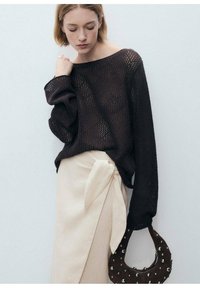 Bruine gebreide trui met wijde mouwen, gecombineerd met een lichtgekleurde wikkelrok met een knoopdetail. Houdt een ronde, met studs versierde handtas vast.