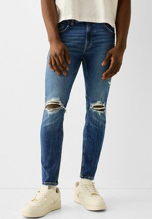 Mand iført slidte blå skinny jeans og hvide sneakers står mod en ensartet lys baggrund.