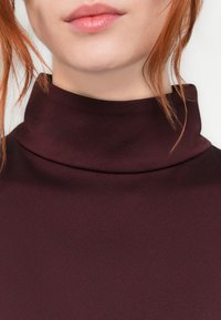 Nærbilde av en person med lys hud og rødt hår som har på seg en mørk burgunder høyhalset genser.