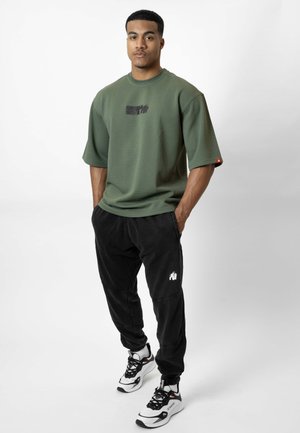 Man staat met handen in de zakken, draagt een oversized groen shirt met korte mouwen, zwarte broek met wit logo en witte zwarte sneakers.