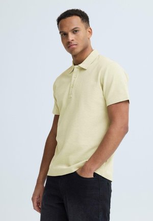 Ung mand stående med den ene hånd i lommen, iført en lysegul kortærmet polo og mørke bukser mod en ensfarvet baggrund.