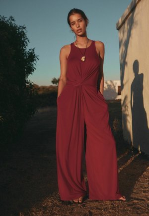 Woman standing outdoors in a sleeveless, wide-leg maroon jumpsuit with hands in pockets, wearing a shell pendant necklace, against a clear sky.