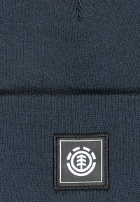 Donkerblauwe gebreide beanie met een gestructureerd oppervlak, voorzien van een rechthoekige zwarte patch met een wit merklogo in het midden.