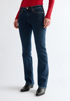 Person iført mørkeblå straight-leg jeans, rød langærmet top og sorte spidse ankelstøvler, der står på en ensfarvet baggrund.