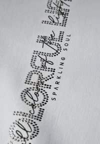 Witte stof met zwarte letters en sprankelende rhinestones in een vloeiende cursieve lettertype, met de tekst "Kleur van het licht" en "SPARKLING SOUL."