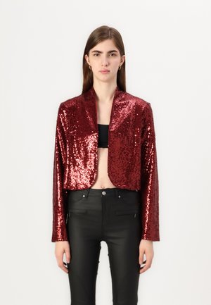 Veste courte avec des sequins rouges, texture lisse et col châle. Portée sur un haut noir et assortie à un pantalon noir ajusté.