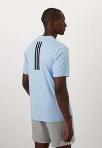 Lyseblå kortermet skjorte med svart tre-stripesdesign på baksiden, laget av et glatt, lett materiale. Modell iført grå korte bukser.