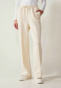 Calças largas em creme com cintura elástica, bolsos laterais e uma textura suave, combinadas com tênis brancos.