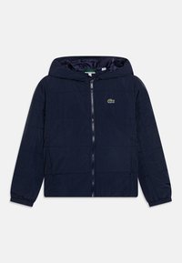 Veste matelassée à capuche bleu marine avec fermeture éclair à l'avant, poignets élastiques et un petit logo vert sur la poitrine. Texture douce avec un design matelassé.