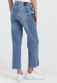 Persona vistiendo jeans de mezclilla de tiro alto en azul claro con piernas rectas y zapatillas Nike Air blancas, de pie frente a un fondo liso.