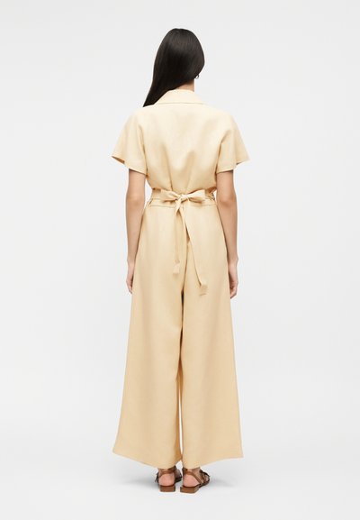 Γυναίκα με μακριά σκουρόχρωμα μαλλιά φοράει μία μπεζ ολόσωμη φόρμα με κοντά μανίκια, φαρδιά μπατζάκια και δεμένο ζωνάρι στην πλάτη, συνδυασμένη με καφέ επίπεδα σανδάλια.