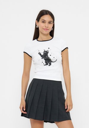 Jeune femme portant un t-shirt blanc ajusté avec un bord noir et un motif chaton noir, associé à une jupe plissée foncée.