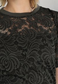 Top de encaje verde oscuro con patrones florales, un cuello redondo con ribete acanalado y mangas cortas, que destaca detalles de textura intrincados.