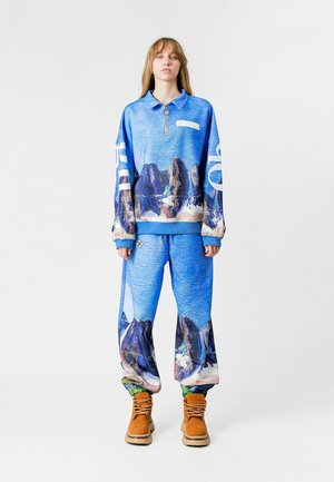 Sudadera azul con un diseño estampado, con media cremallera y mangas anchas, combinada con pantalones a juego que presentan un diseño de montañas y detalles de cordón. Botas marrones.