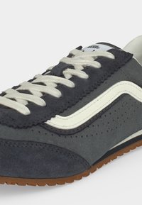 Σκούρο γκρι αθλητικό παπούτσι Vans με λευκή πλαϊνή λωρίδα, λευκά κορδόνια, διάτρητες λεπτομέρειες και σόλα από καουτσούκ χρώματος καφέ.