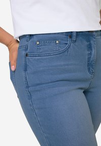 Jeans de mezclilla azul con un ajuste slim, con una cintura de altura media, dos bolsillos frontales y detalles en botones de metal. Textura suave, desvanecido mínimo.