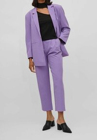 Ensemble costume violet comprenant un long blazer et un pantalon fuselé. Haut noir avec un col ras du cou. Mules noires à talons épais complètent la tenue.