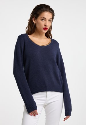Frau mit dunklem welligem Haar, die einen marineblauen Pullover mit goldener Kettenverzierung und weiße Hosen trägt, posiert vor einem schlichten hellen Hintergrund.