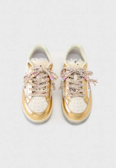 Λευκά και χρυσά sneakers χαμηλού ύφους με πλεκτά κορδόνια που διαθέτουν πολύχρωμες χάντρες σε απλό ανοιχτό γκρι φόντο.