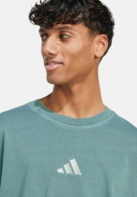 Tyrkysové bavlněné tričko s kulatým výstřihem a bílým logem Adidas s třemi pruhy na levé části hrudi. Hladká textura.